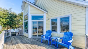A wooden deck with blue Adirondack chairs and a table, a sliding glass door, and a yellow building with white trim.