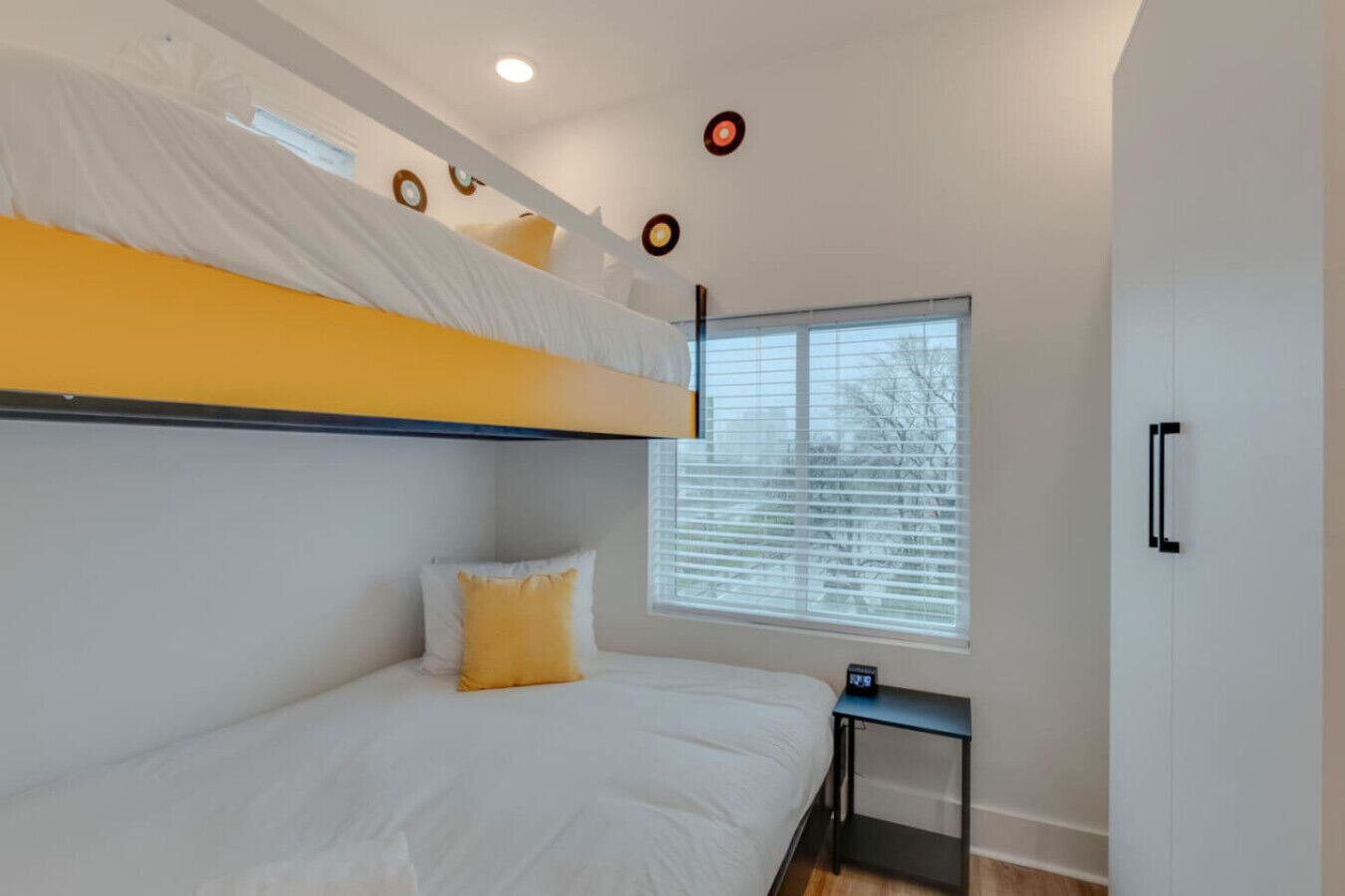 A modern bedroom with a bunk bed, a bedside table with a clock, and a window with blinds.