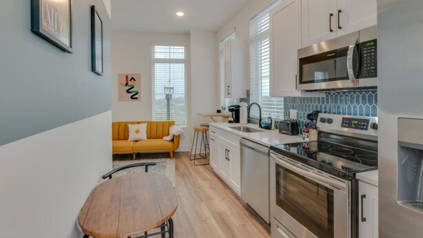 A modern kitchen with white cabinets, a microwave, an oven, and a sink. There is a yellow sofa and a wooden table in the adjacent living area.