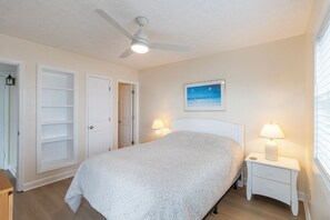 Une chambre à coucher comprenant un lit, des tables de chevet, un ventilateur de plafond et une fenêtre avec des stores.