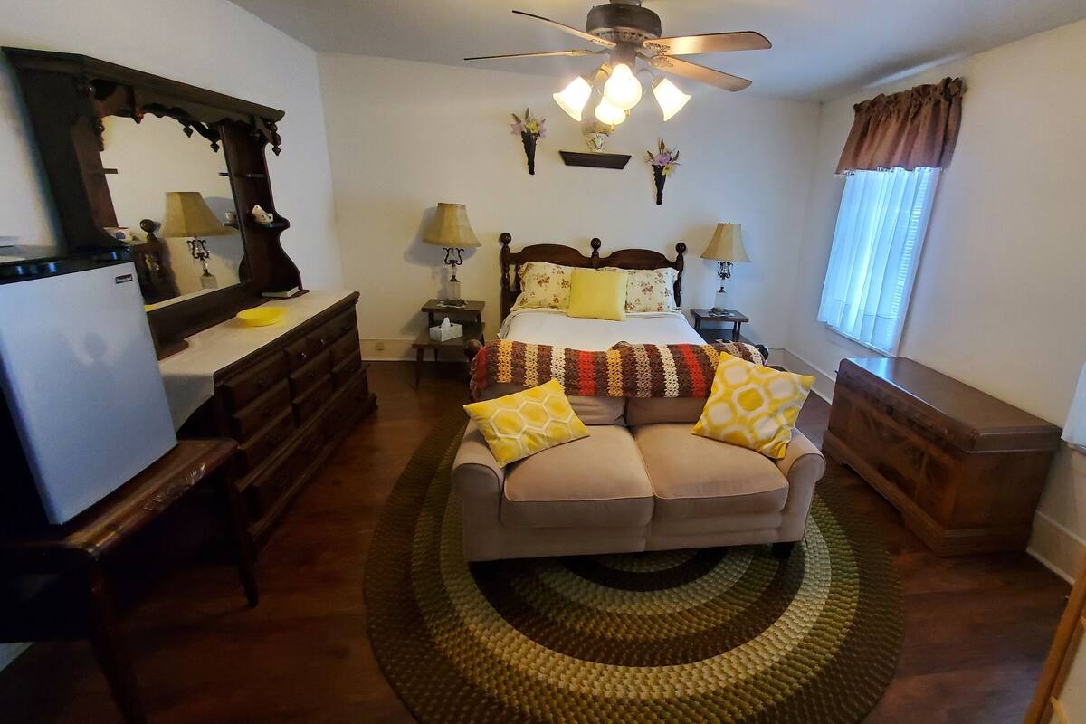 Un dormitorio con cama, mesitas de noche, cómoda, espejo, ventilador de techo y ventana con cortinas.