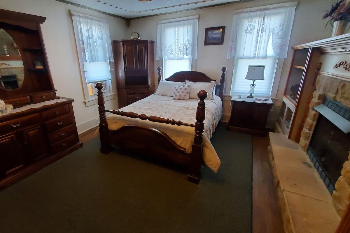 Un dormitorio tradicional con una cama de madera con dosel, un aparador, un espejo, una chimenea y cortinas de encaje en las ventanas.