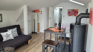 Een moderne woonkamer met een bank, een tafel, een klein krukje, een glazen salontafel, een zwarte heater en een kast met hangende kleding.