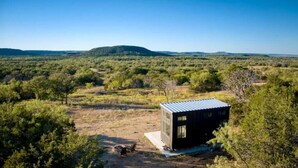 A small, dark-colored cabin in a natural landscape with sparse vegetation and distant hills. - Tiny Home Near Palo Pinto Mountains State Park + Scenic Views + Fire pit + Grill (Ranger)