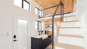 A modern kitchen with a central island, a staircase, and a view of the outdoors. - Tiny Home Near Palo Pinto Mountains State Park + Scenic Views + Fire pit + Grill (Ranger)