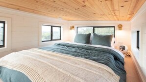 A bedroom with a wooden ceiling, large windows, a bed with a green and white duvet, and a bedside table with a lamp. - Tiny Home Near Palo Pinto Mountains State Park + Scenic Views + Fire pit + Grill (Ranger)