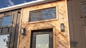 Un edificio moderno con fachada de madera, una puerta central y un pequeño espacio al aire libre con mesas y sillas.