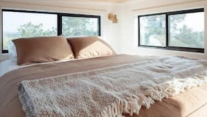 Una camera da letto con soffitto in legno, ampie finestre, un letto con biancheria beige e una coperta in maglia.
