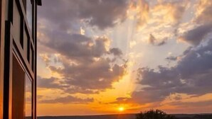 Une vue sur le coucher de soleil, prise depuis une fenêtre donnant sur un espace verdoyant avec quelques arbres et un bâtiment au premier plan.