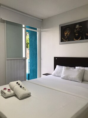 A hotel room with a bed, white pillows, a black headboard, a nightstand, a towel with a bottle, and a painting on the wall. - HOTEL SAN ALEJO (Tulua)