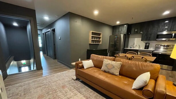 Una sala moderna con un sofá de cuero marrón, un comedor con muebles de madera y una cocina con gabinetes oscuros y electrodomésticos de acero inoxidable.