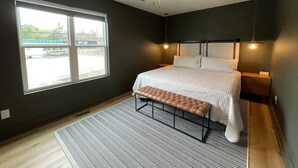 Un dormitorio con cama, banca, ventilador de techo, ventana y mesita de noche.