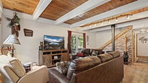 A cozy living room with a brown leather sofa, a white armchair, and a wooden entertainment unit. A large TV is mounted on the wall, and there's a staircase leading to another level.