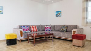 Un salon avec un canapé gris, des coussins colorés, une table basse en bois et deux poufs, un jaune et un rouge.