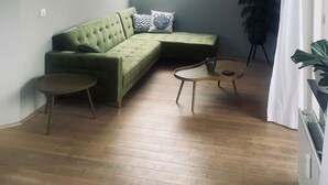 A modern living room with a green sectional sofa, two small side tables, a large window with natural light, and a potted plant. - Main Street Beach apartment 150 meters from the beach  (Noordwijk)