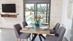 Een eethoek met een tafel voor vier personen, grijze bekleden stoelen, een houten tafel en uitzicht op een zwembad en groen buiten.