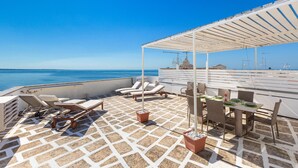 Una terraza al aire libre iluminada por el sol, con sillones, una mesa y una pérgola con vistas al mar.