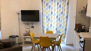 Un salon moderne avec un canapé, une table à manger entourée de chaises jaunes, un téléviseur à écran plat fixé au mur et un coin cuisine équipé d’une cuisinière et de placards.