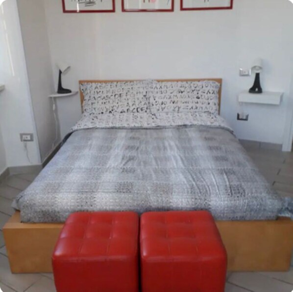 Un dormitorio con una cama, dos otomanes rojos, una mesita de noche, una lámpara y cuadros en la pared.