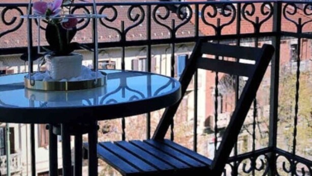 Un balcon avec une table ronde et une chaise, un pot de fleurs, et une vue sur des bâtiments et des montagnes en arrière-plan.
