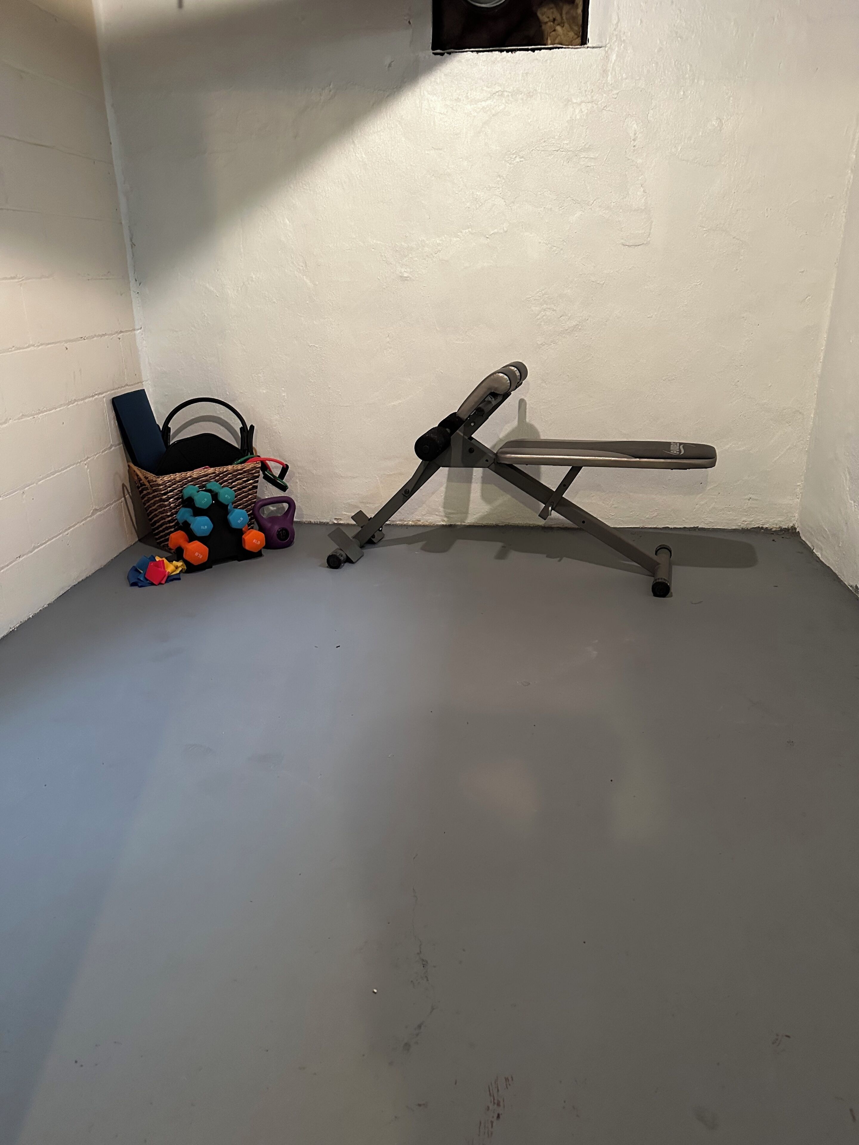 Canto de um quarto com parede branca, luminária de chão e cesta de vime com halteres e tapete de yoga.