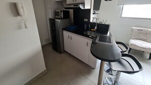 A compact kitchen with white cabinets, a stainless steel refrigerator, a microwave, and a sink. There's a small dining table with chairs, a window providing natural light, and a ceiling-mounted light fixture. - 506 Moderno Aparta-Suite en Versalles Tipo Loft - Cali Tower Suites & Lofts (Cali)