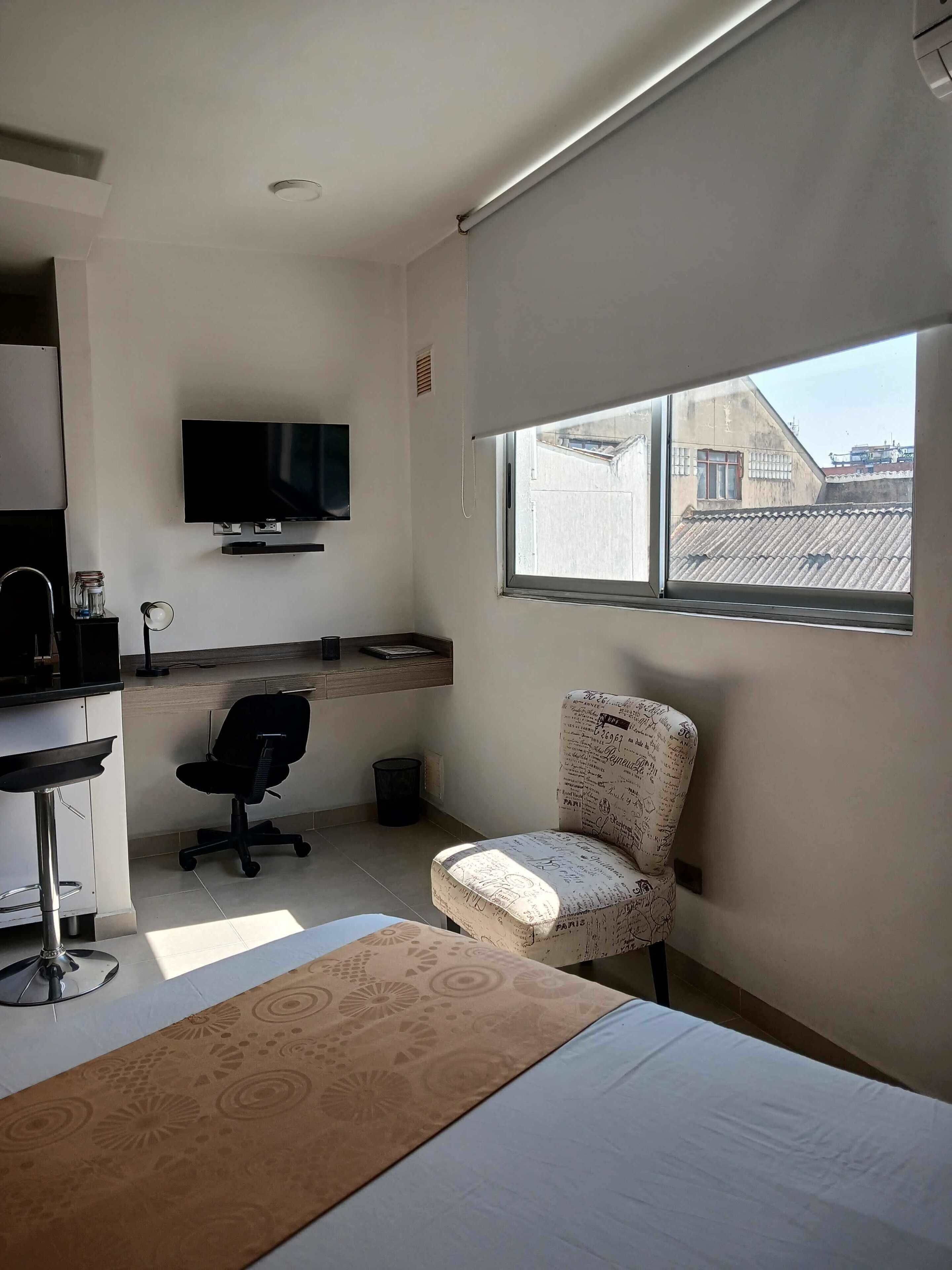 Una habitación moderna con cama, escritorio con silla, televisor montado en la pared y ventana con vista a edificios.
