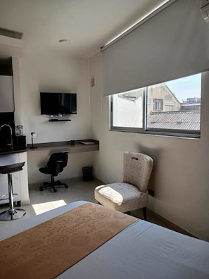 A modern room with a bed, a desk with a chair, a television mounted on the wall, and a window with a view of buildings. - 506 Moderno Aparta-Suite en Versalles Tipo Loft - Cali Tower Suites & Lofts (Cali)