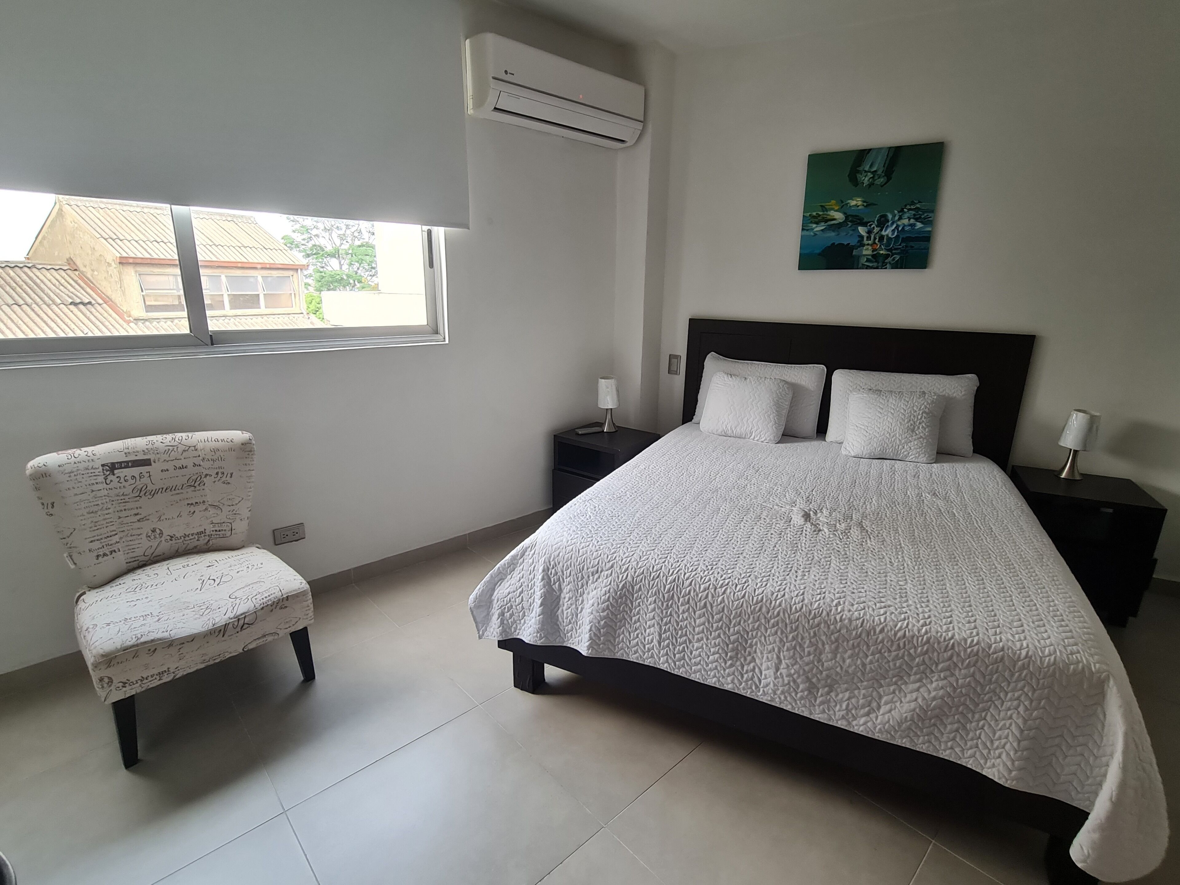 Une chambre à coucher avec un lit, une chaise, une table de chevet, une lampe et un tableau au mur.