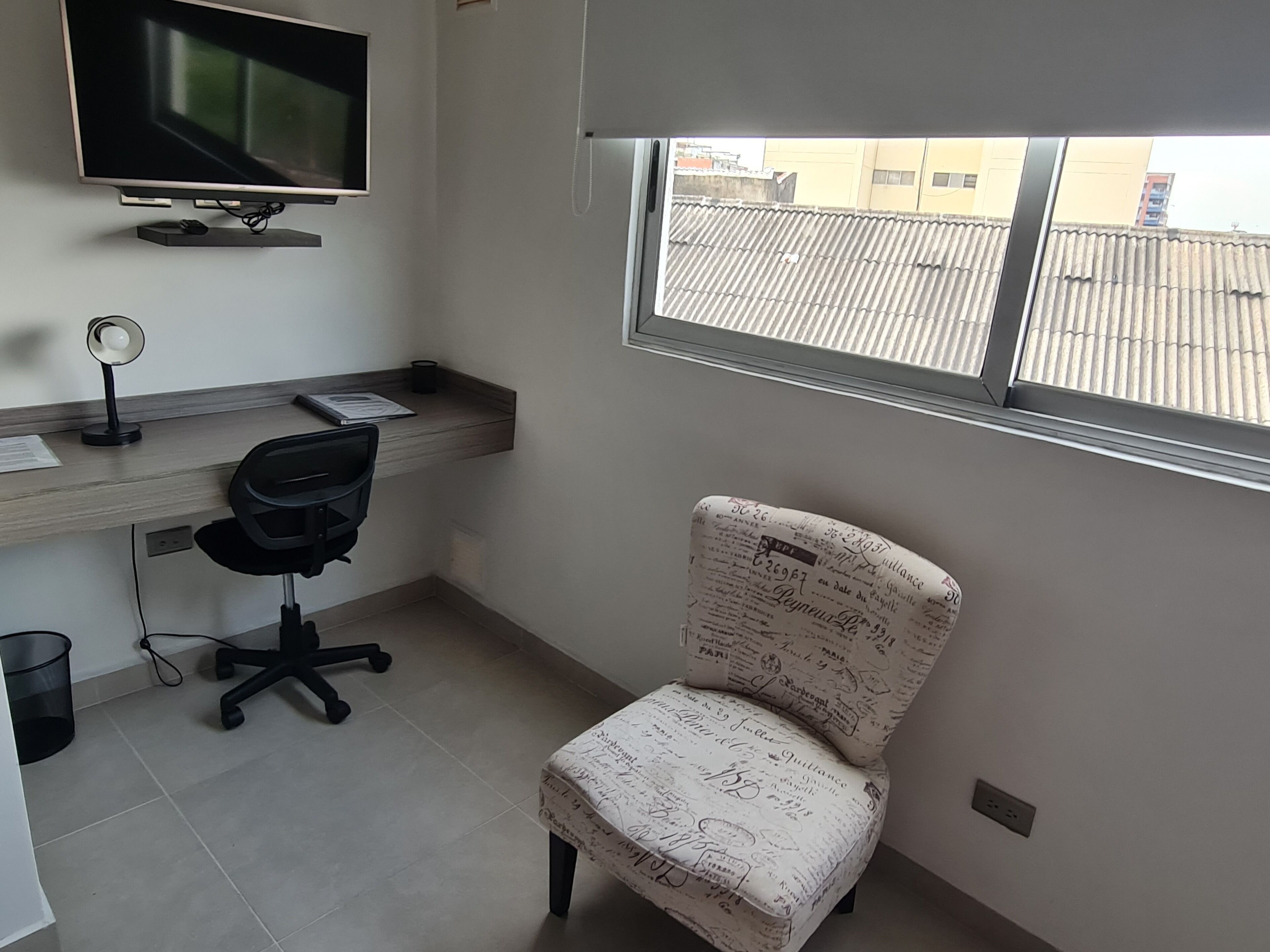 A small office with a desk, chair, TV, and a window with a view of buildings.