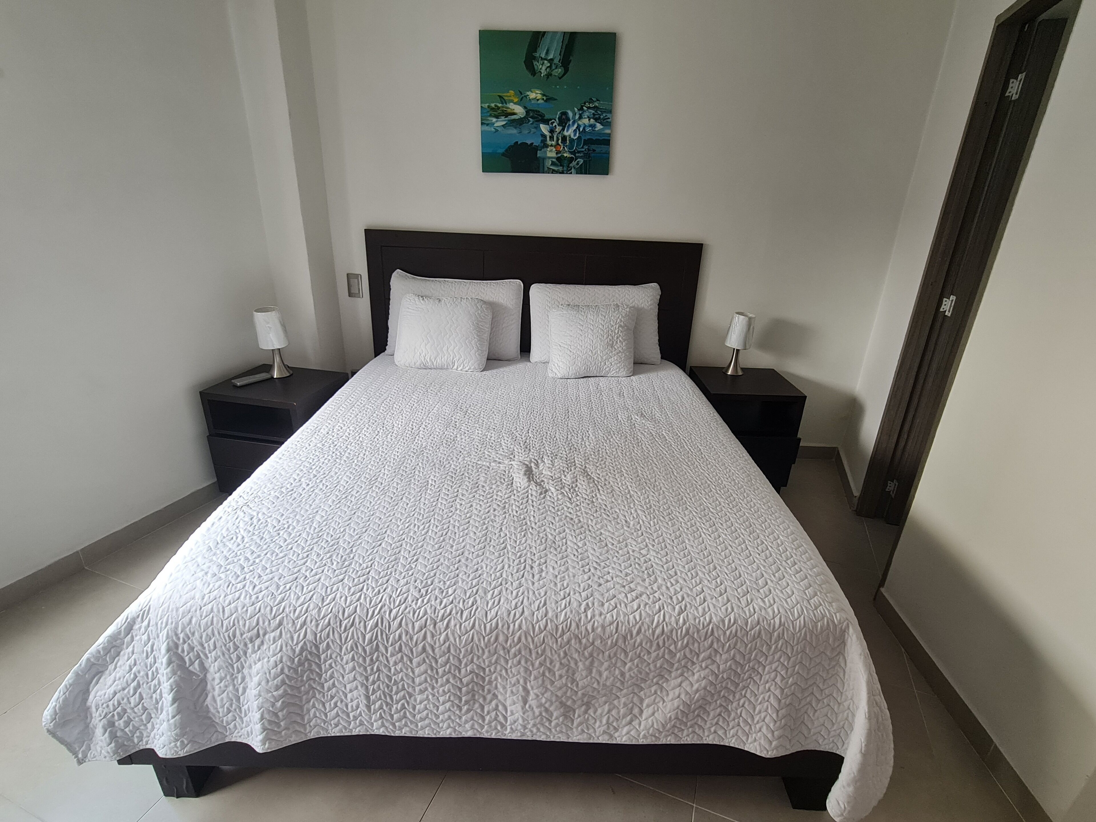 A neatly made bed with a white quilt, two bedside tables with lamps, and a painting on the wall.
