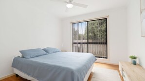 Een slaapkamer met een bed, een plafondventilator, een raam met uitzicht op groen en een kleine tafel met een plant.