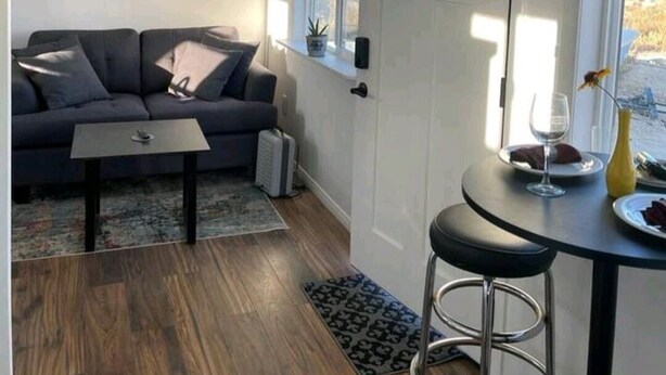 Ein kompakter Wohnbereich mit einem dunkelgrauen Sofa, einem kleinen Tisch, einem Barhocker und einem Deckenventilator.