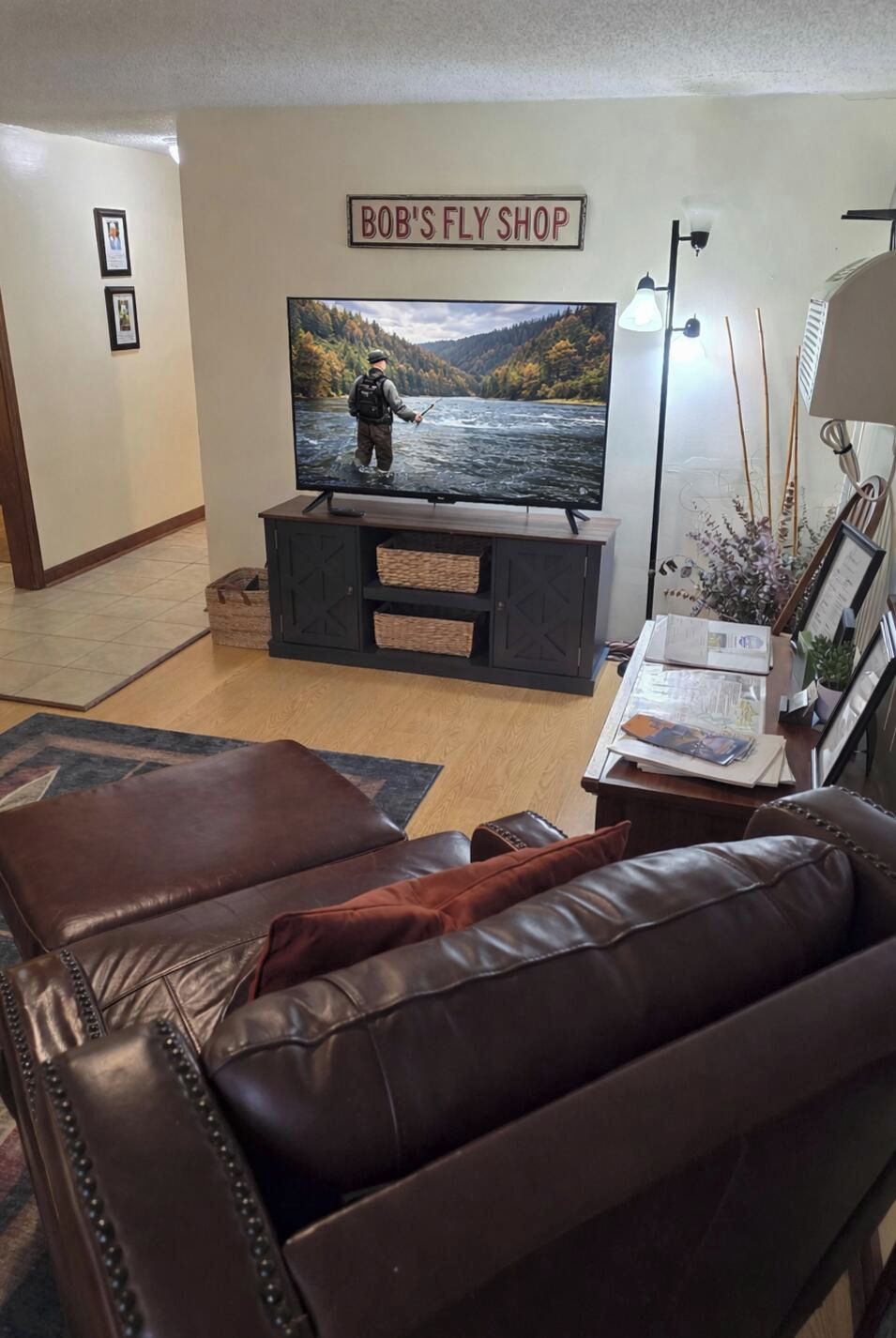 Ein Wohnzimmer mit einem Fernseher, der eine Szene beim Angeln zeigt, einer dunklen Holz-Unterhaltungseinrichtung, einem braunen Ledersofa und einem gerahmten Schild über dem Fernseher mit der Aufschrift 'BOB'S FLY SHOP'.