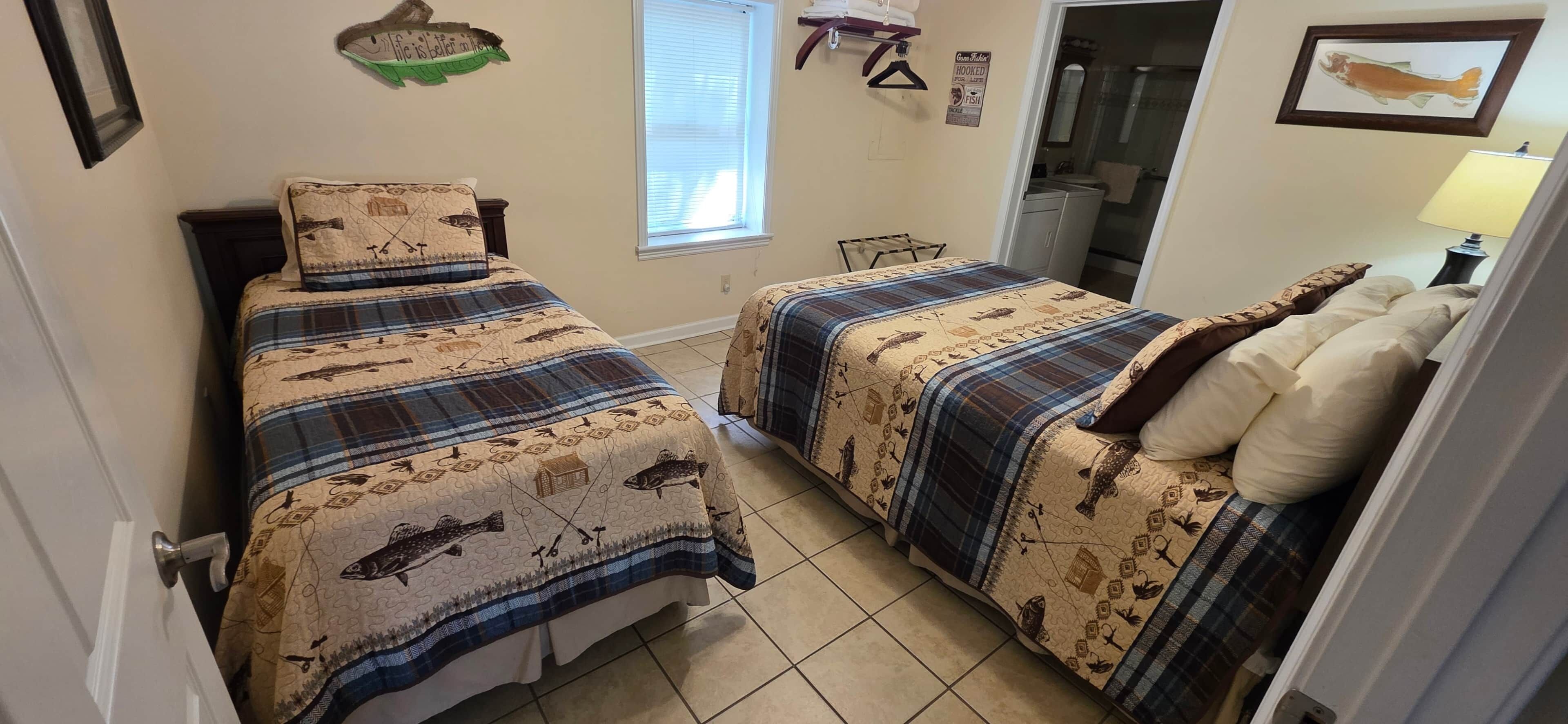 Une petite chambre avec deux lits, chacun recouvert d’une couvre-lit à motifs de poissons, une fenêtre avec des stores, un porte-serviettes et un tableau représentant des poissons accroché au mur.