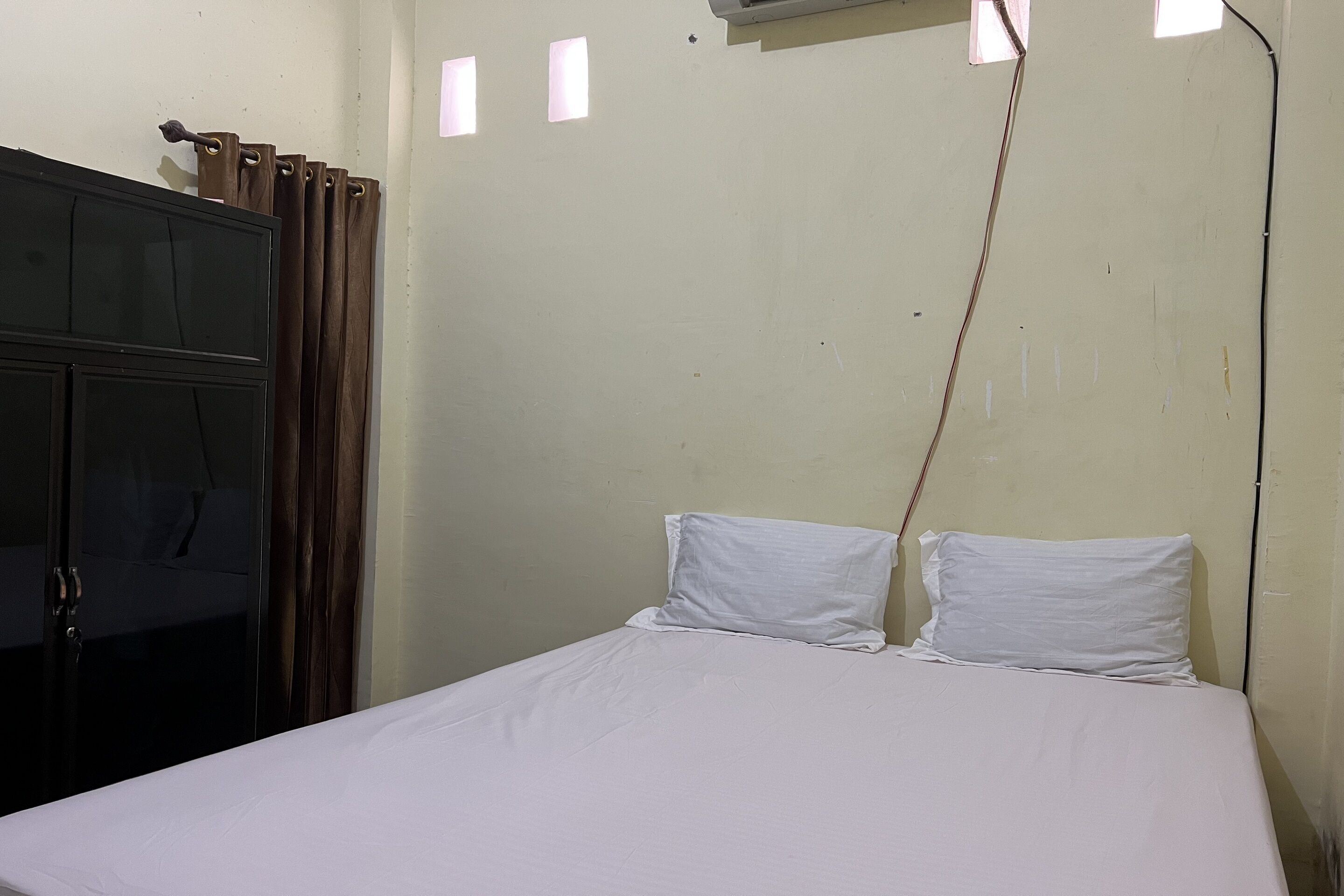 A single bed with white bedding in a room with a plain wall, a window with curtains, and an air conditioning unit mounted on the wall.