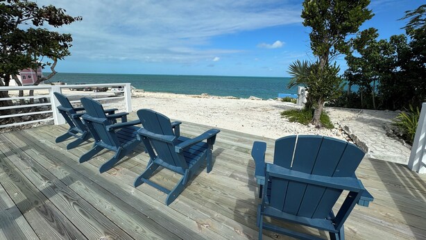 A wooden deck with blue Adirondack chairs facing a beach.