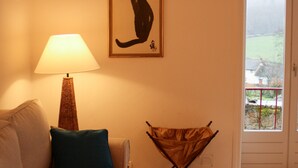 Een woonkamer met een bank, een lamp, een foto van een kat in een lijst en een raam met uitzicht.