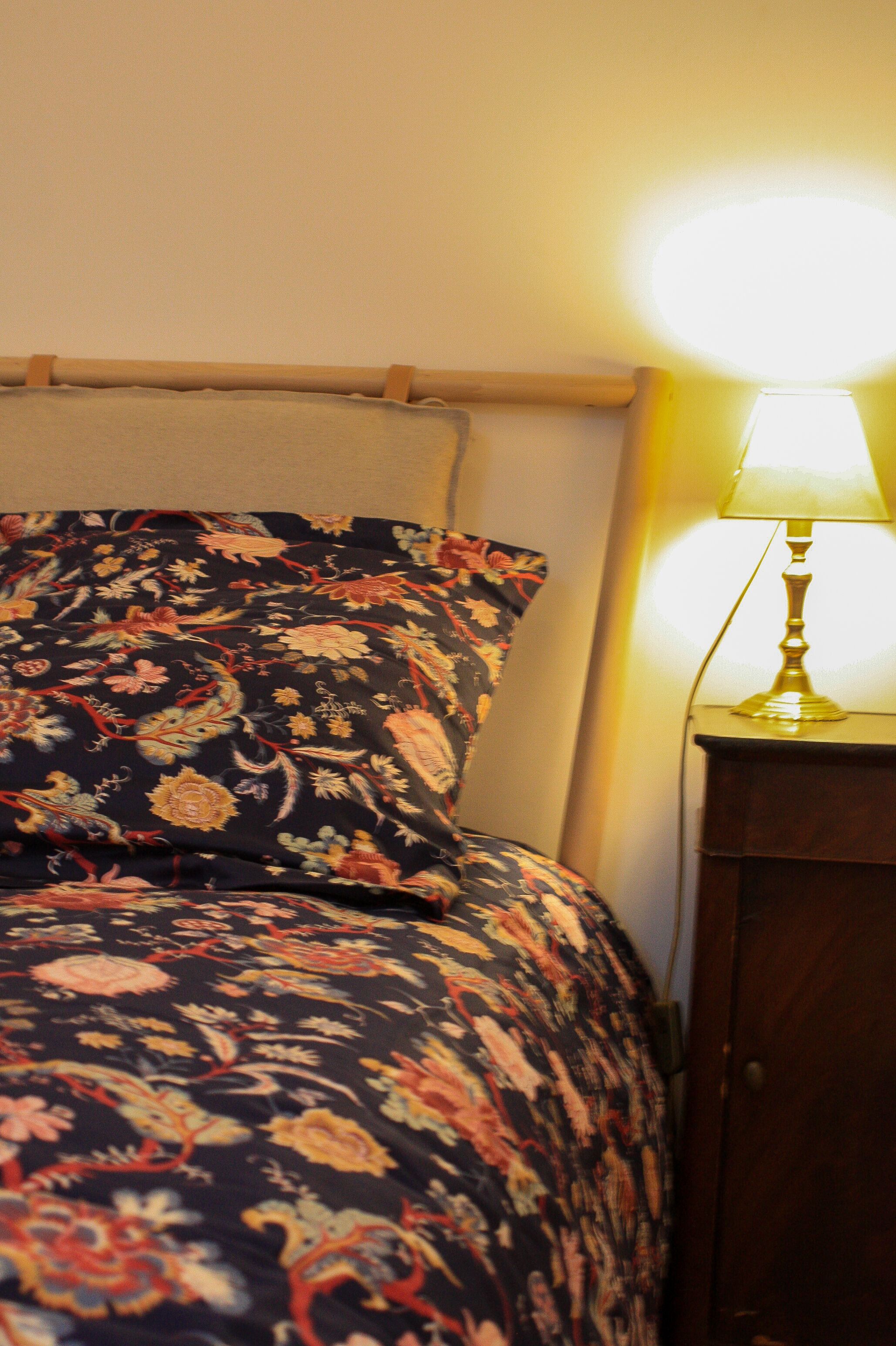 A bed with a floral-patterned bedspread and pillows, a wooden headboard, and a bedside table with a lamp.