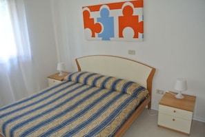 Un dormitorio con una cama, dos mesitas de noche, una mesita auxiliar, una obra de arte en la pared y una ventana con cortinas.