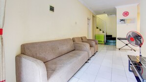 Una habitación con un sofá color beige, un ventilador de pie y una escalera al fondo.