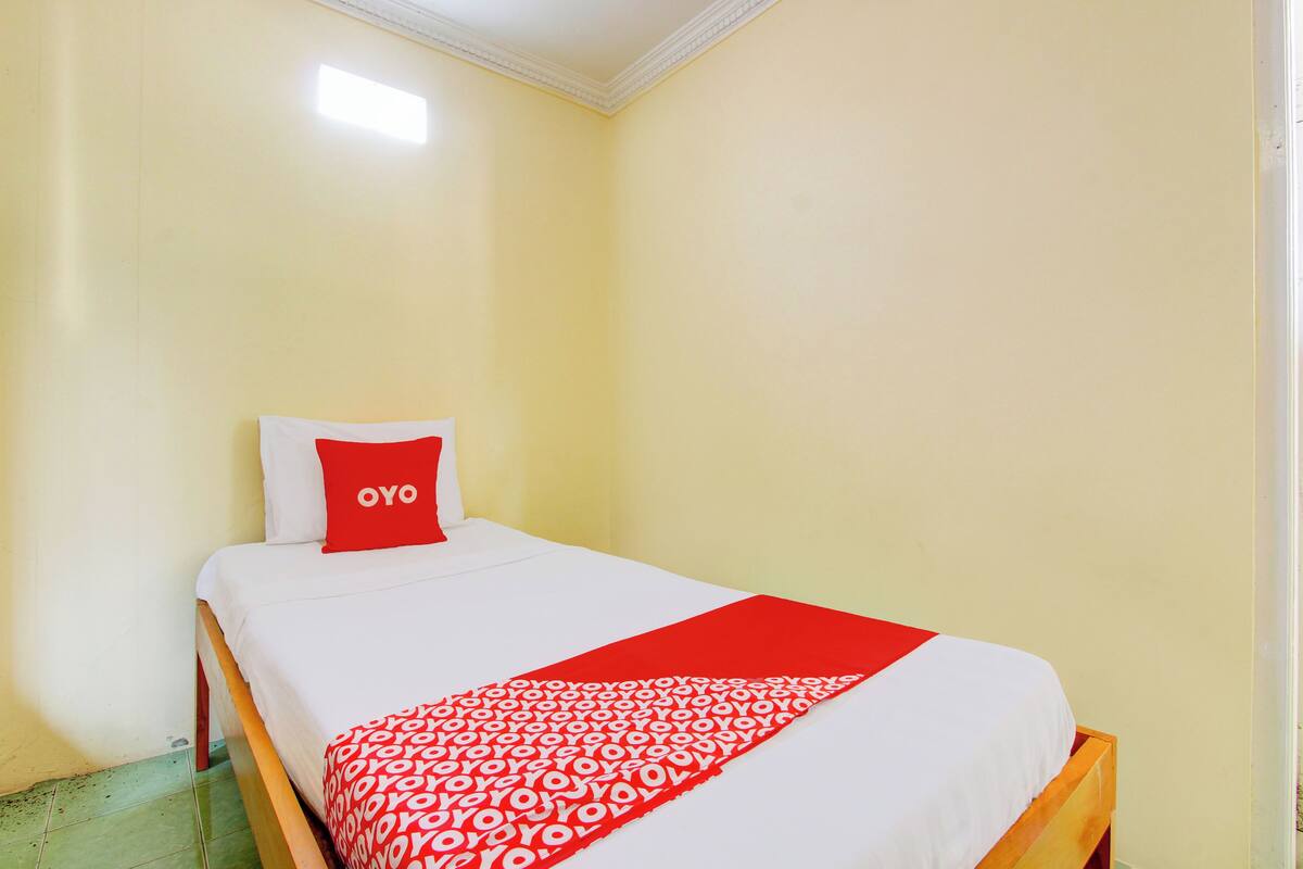 A single bed with a white and red bedspread, a red pillow with the word "OYO" on it, and a wooden headboard.