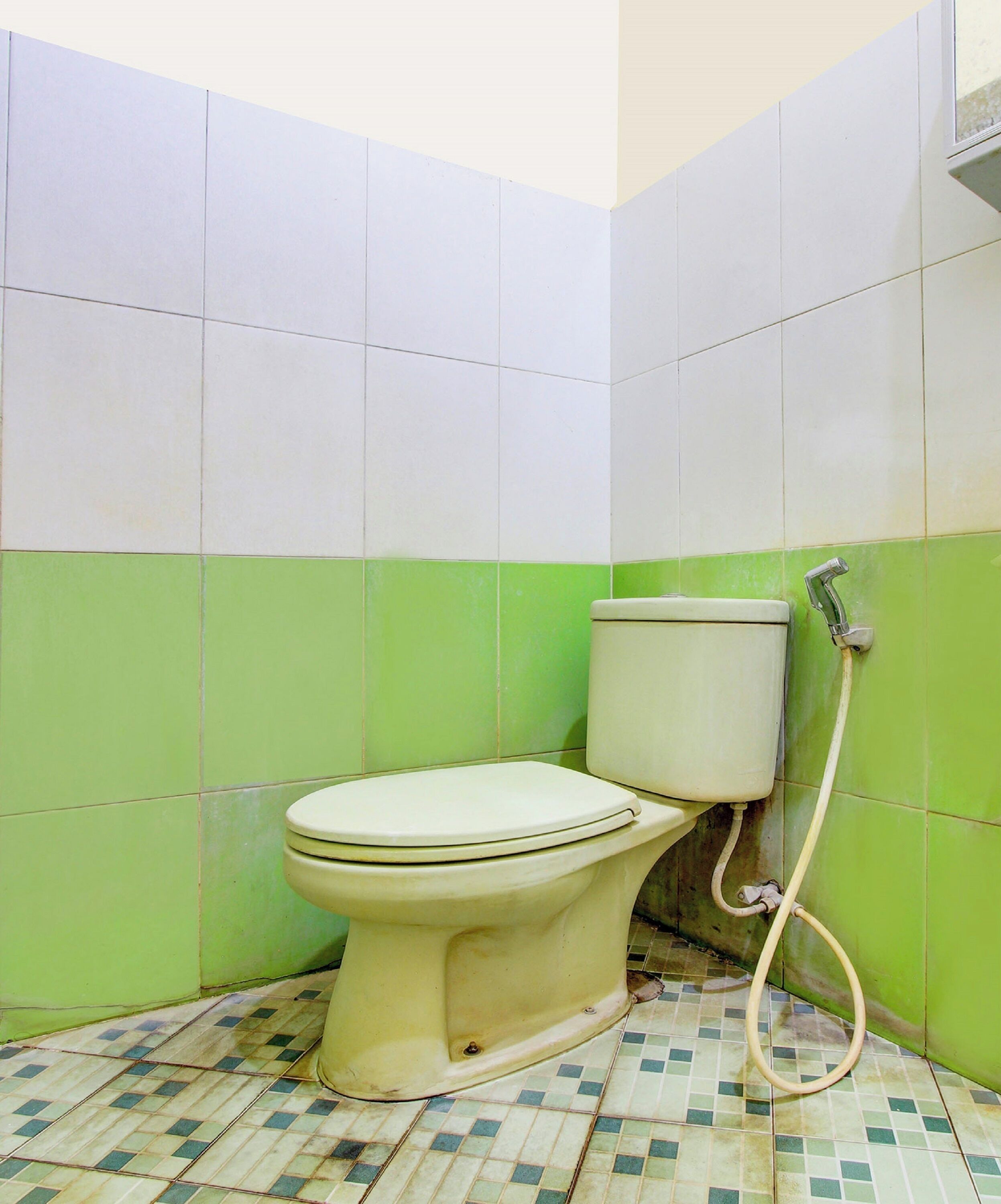 Un baño pequeño y sencillo, con inodoro, paredes de azulejos blancos y verdes, y suelo de baldosas.