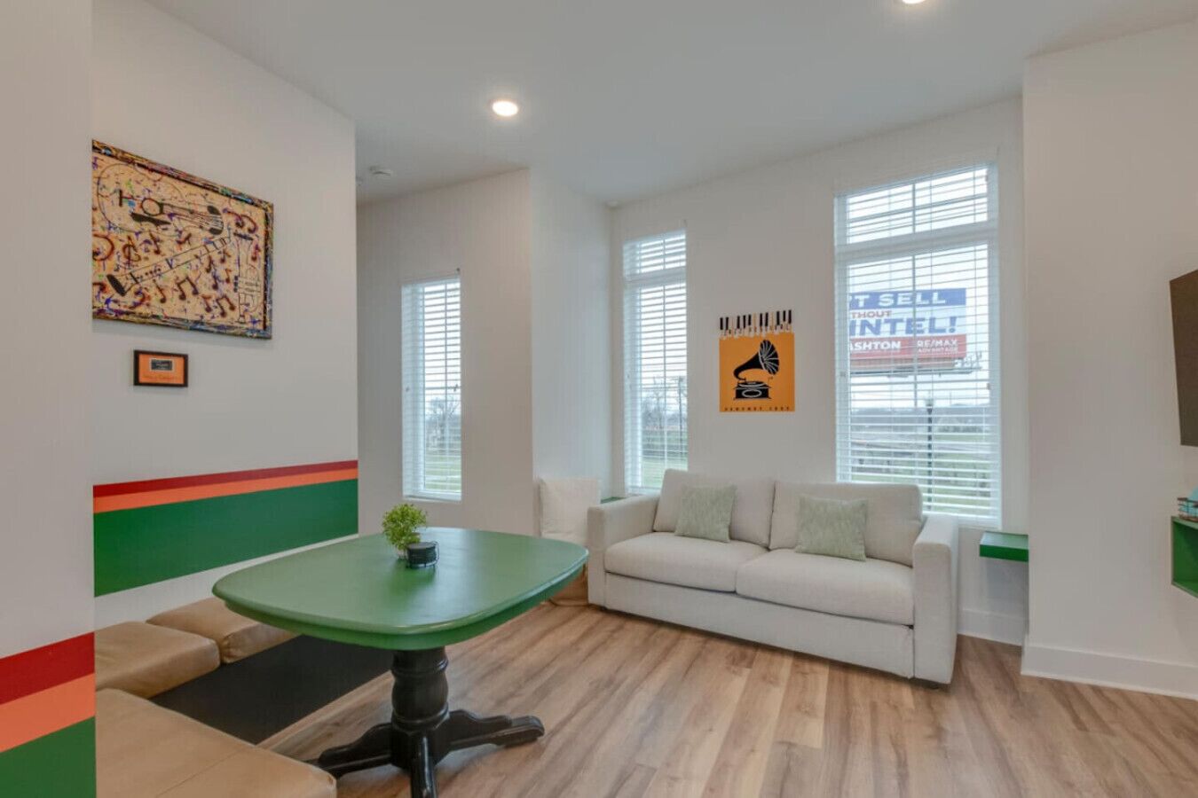 A modern living room with a green table, a white sofa, and a large window with blinds.