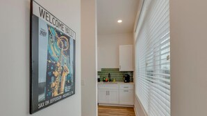 Un pasillo con un cartel en la pared, una zona de cocina con armarios y salpicadero de azulejos, y una ventana con persianas.