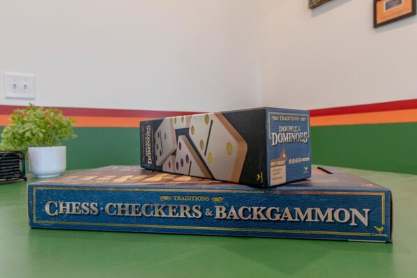 A chess set titled "Chess Checkers & Backgammon" on a table with a plant and a colorful wall in the background.