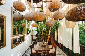 A dining area with wicker hanging lamps, wooden furniture, and a view of lush greenery outside. - La Ferme Medina (Marrakech)