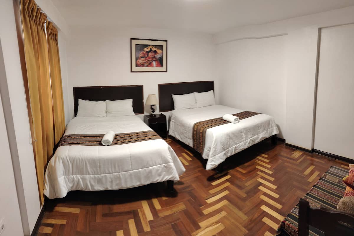 A hotel room with two beds, a wooden herringbone floor, a nightstand with a lamp, and a framed picture on the wall.