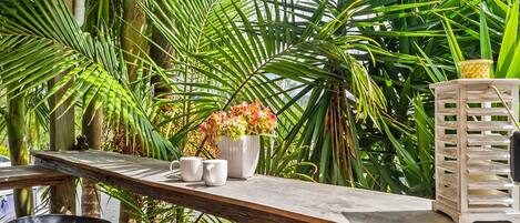 A wooden table with two white mugs and a vase of flowers, surrounded by lush green foliage.