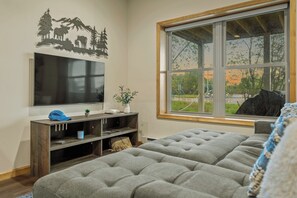 A bedroom with a large window, a flat-screen TV mounted on the wall, a wooden console table, and a tufted grey bed. - Pocono Paradise by Avantstay Lakeside Retreat, Hot Tub, Game Room, Near Beach (Albrightsville)